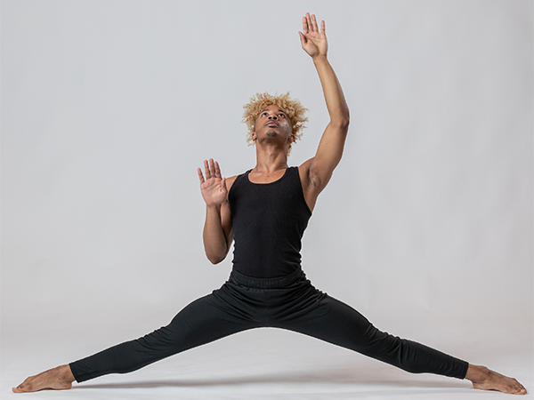 A college student poses in a dancing posture, legs stretched wide and arm extended upwards, while wearing black dancing attire.